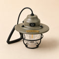 An olive green and black Staurk portable lantern with a clear glass body, black wire cage, and a gold-colored brand plate, set against a solid beige background.